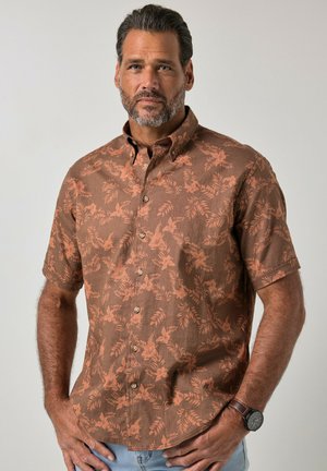 Mann mit salt-and-pepper Haar und Bart, der ein braunes, kurzärmeliges Hemd mit orangefarbenen Blumenmustern und eine Armbanduhr trägt.
