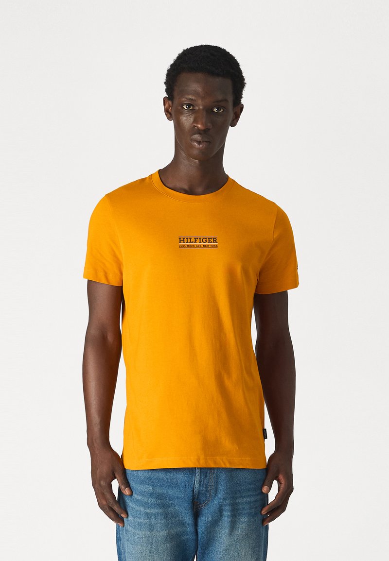 T-shirt di cotone arancione con maniche corte, collo rotondo e logo "HILFIGER" stampato in nero. Indossato con jeans in denim blu. Design semplice, senza fantasie.