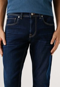 Tmavomodré džínsy so slim fit strihom, s svetlomodrým prešívaním pozdĺž vreciek a švov, a s gombíkovým zapínaním v páse.