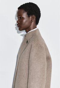Beżowy wełniany płaszcz z wysokim kołnierzem, z widocznym guzikiem i prostymi liniami. Gładka faktura, dopasowany krój i minimalistyczny design.
