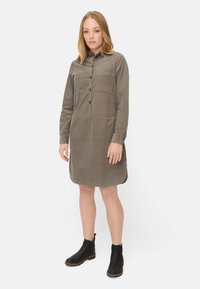 Robe chemise en velours côtelé vert olive avec des manches longues, fermeture à boutons sur le devant, deux poches poitrine et un ourlet arrondi, accompagnée de bottines noires.