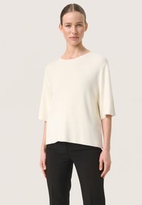 Woman with pulled-back hair wearing a loose, short-sleeve white top and black pants, standing against a plain light background.