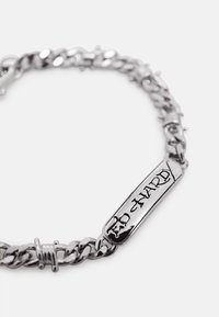 Strieborný náramok s reťazou s obdĺžnikovou platničkou, na ktorej je rytý nápis "ED HARDY" v štýlovom písme na bielom pozadí.