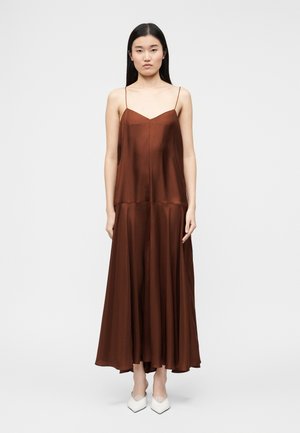 Mujer con cabello largo y negro, vistiendo un vestido largo de satén marrón sin mangas y zapatos blancos con punta, de pie frente a un fondo blanco liso.