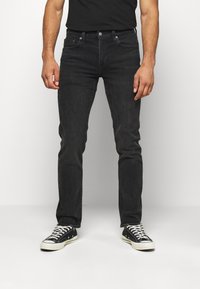 Jean en denim noir à coupe ajustée, doté d'un design classique à cinq poches, d'accessoires métalliques, et associé à des baskets noires et blanches.