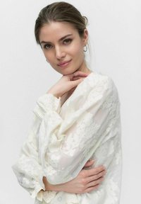 Hvid blonder bluse med lange ærmer og elastiske manchetter, der har et gennemsigtigt materiale og delikate blomsterbroderi detaljer.