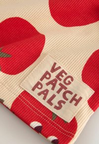 Béžová žebrovaná látka s velkými červenými potisky rajčat a přišitou obdélníkovou cedulkou s nápisem "VEG PATCH PALS" tučnými písmeny.
