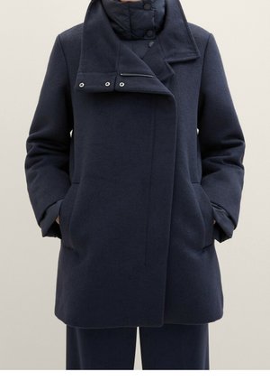 Femme portant un manteau en laine bleu marine foncé avec une fermeture frontale cachée, des poches latérales et un col et des poignets intérieurs matelassés et superposés.