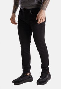 Pantaloni neri in misto cotone con vestibilità slim, dotati di chiusura frontale con bottone e tasche laterali. Abbinati a scarpe sportive nere.