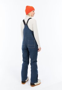 Vrouw in blauwe denim overall, beige geribbelde coltrui, feloranje beanie en witte schoenen staat zijwaarts gekeerd voor een witte achtergrond.