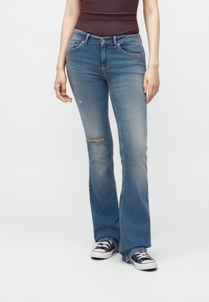 Person iført blå udsvingede jeans med let slid på knæet og sorte lave sneakers, står mod en ensfarvet baggrund.