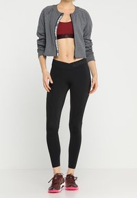 Giacca grigia cropped con logo, reggiseno sportivo bordeaux, leggings neri con texture e sneaker rosso scuro con suole rosa.