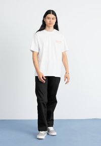 Witte t-shirt met korte mouwen met een oranje logo, gecombineerd met zwarte broek en grijze schoenen, op een lichte achtergrond.