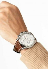 Reloj de cuero marrón con caja de acero inoxidable plateado, esfera blanca y características de cronógrafo. Textura de hilo visible en el suéter de la muñeca.