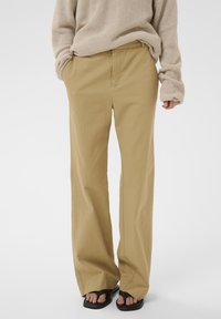 Personne portant un pull en maille beige et un pantalon ample marron clair, mains dans les poches, debout en sandales noires sur fond blanc.