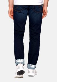 Jeans de mezclilla azul oscuro con costuras naranjas contrastantes, combinados con zapatillas deportivas blancas que tienen un puño de tobillo gris claro etiquetado como "CAMP DAVID."