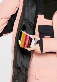 Ski goggles met een wit frame en kleurrijke spiegelende lenzen vastgehouden in een hand. De persoon draagt een roze en zwarte jas met zichtbare zakken.