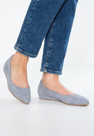 Fødder i blå jeans iført lysegrå ruskind kilehælssko, der står på en hvid overflade.
