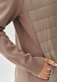 Maglione marrone con una sezione toracica trapuntata e imbottita. Texture morbida con polsini a coste e una mano in una tasca laterale.
