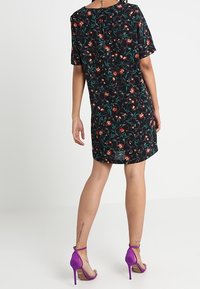 Robe noire à fleurs avec manches courtes, ornée de motifs rouges et verts. Associée à des sandales à talons hauts violettes.