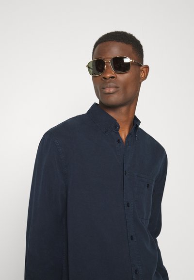 Camicia azzurra con bottoni, dotata di tasca a toppa, indossata sotto occhiali da sole scuri. La camicia presenta un colletto classico e una texture liscia.