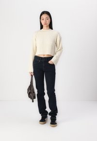 Cream ribbed crop sweater with long sleeves, paired with dark denim jeans and black sneakers. Model holds a small brown bag.
