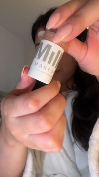 Flasche von Milk Makeup mit einem halbtransparenten Deckel, weißem und grauem Etikett, gehalten in einer Hand mit zartrosafarbenen Nägeln. Mattes Finish, kompaktes Design.