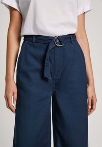 Navy blauwe hoge taillebroeken met een brede pijp, voorzien van een stoffen riem met een metalen ring en zijzakken. Soepele katoen textuur.
