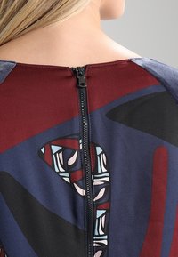 Farbiger Kleiderrücken mit einem schwarzen Reißverschluss; zeigt kastanienbraune, blaue und abstrakte geometrische Muster in Beige und Türkis. Glatte Stofftextur.
