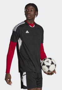 Schwarzes Fußballtrikot mit strukturiertem Muster, weißen Akzenten und drei weißen Streifen. Rote lange Ärmel unter dem Trikot. Hält einen schwarz-weißen Fußball.