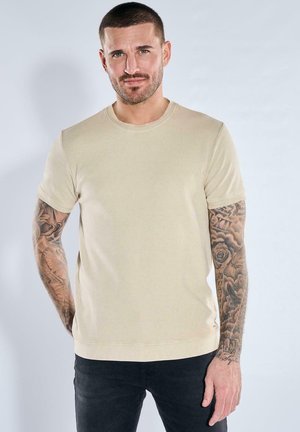 Mann mit kurzen Haaren und Bart, trägt ein beiges Kurzarmhemd und schwarze Hose, zeigt detaillierte Tattoos auf beiden Armen vor hellem Hintergrund.