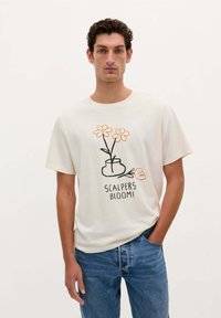 Witte katoen T-shirt met een florale graphic in oranje en zwart, met de tekst "SCALPERS BLOOM!" onder het ontwerp. Gedragen met blauwe spijkerbroeken.