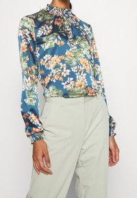 Blouse fleurie avec un fond turquoise, présentant des fleurs orange et blanches. Manches longues avec poignets élastiques et encolure froncée.