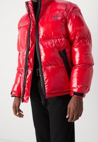 Rote Steppjacke mit glänzendem Finish, ausgestattet mit Reißverschluss, Seitentaschen und schwarzen Akzenten. Logo auf der linken Brustseite sichtbar.