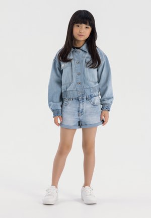 Veste en denim bleu clair avec taille élastique, associée à un short bleu clair retroussé et des baskets blanches, sur un fond neutre.