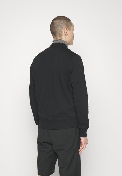 Svart hettegenser med ståkrage, ribbestrikkede mansjetter og nederkant, vist fra baksiden. Stoffet ser glatt ut og er tettsittende i midjen.