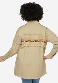 Giacca beige chiaro con schiena testurizzata caratterizzata da pizzo, paillettes dorate e dettagli con frange, abbinata a maniche arrotolate.