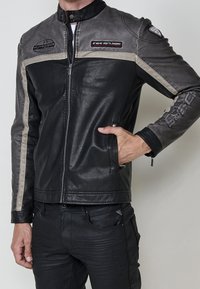 Homme portant une veste de motard en cuir noire et grise avec des patchs et des fermetures éclair, main dans la poche, associée à un pantalon noir.