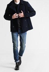 Marineblauwe wollen peacoat met dubbelrijige knopen, gedragen over een donker sweater en lichtblauwe spijkerbroek, gecombineerd met zwarte enkellaarzen.