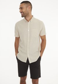 Camicia beige con bottoni e maniche corte, dotata di tasca, realizzata in tessuto strutturato; indossata con pantaloni corti scuri, in piedi davanti a uno sfondo neutro.