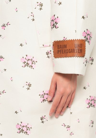 Fehér szövet virágmintás rózsaszín és barna hímzéssel, amelyen egy texturált barna címke található, rajta "BAUM UND PFERDGARTEN" felirattal az ujján.
