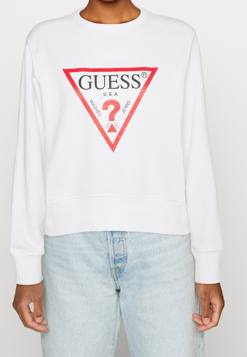Persona che indossa una felpa bianca con il logo triangolare rosso GUESS e jeans azzurri chiari, in piedi davanti a uno sfondo semplice.