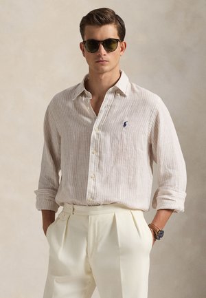 Jasnobeżowa lniana koszula w paski z kołnierzykiem na guziki, zestawiona z kremowymi plisowanymi spodniami. Okulary przeciwsłoneczne z ciemnymi szkłami dopełniają stylizację.