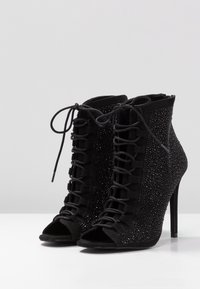 Zwarte enkellaarsjes met hoge hak en open neus, voorzien van een veterluiting aan de voorkant en een gestructureerde, glinsterende stof aan de zijkanten.
