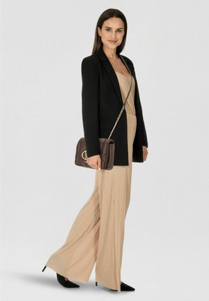 Mujer de pie, vistiendo un mono plisado beige con blazer negro, tacones negros puntiagudos y llevando un bolso de hombro marrón oscuro con cadena dorada.