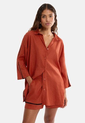 Kvinde iført en løstsiddende, rustfarvet skjorte med knapper og matchende shorts med sort kant, med hånden i lommen, vendt fremad mod en ensfarvet baggrund.