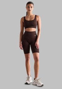 Smeđa rebrovana sportska grudnjak i set visokih biciklističkih kratkih hlača. Ima prilagođeni kroj i bezšavnu izvedbu. Uparen s bijelim sportskim tenisicama.