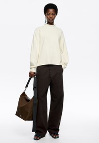 Cremig strukturierter Pullover, lockere dunkelbraune Hose, trägt eine braune Tasche mit grauem Träger und blaue gemusterte Sandalen.