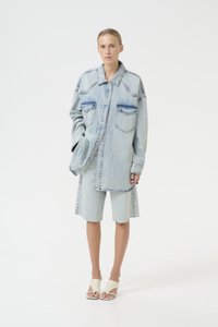 Giacca oversized in denim azzurro chiaro con due tasche frontali, abbinata a pantaloni corti in denim azzurro chiaro. Il modello indossa sandali in crema.