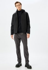 Chaqueta negra con dos bolsillos en el pecho, cierre de cremallera y capucha ajustable; combinada con pantalones grises y botas negras de suela gruesa. Acabado mate, diseño casual.